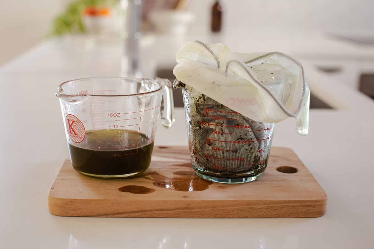 A jug on the left is filled with strained, infused plantain oil, while a jug on the left contains the used herb in a muslin cloth ready to be composted.