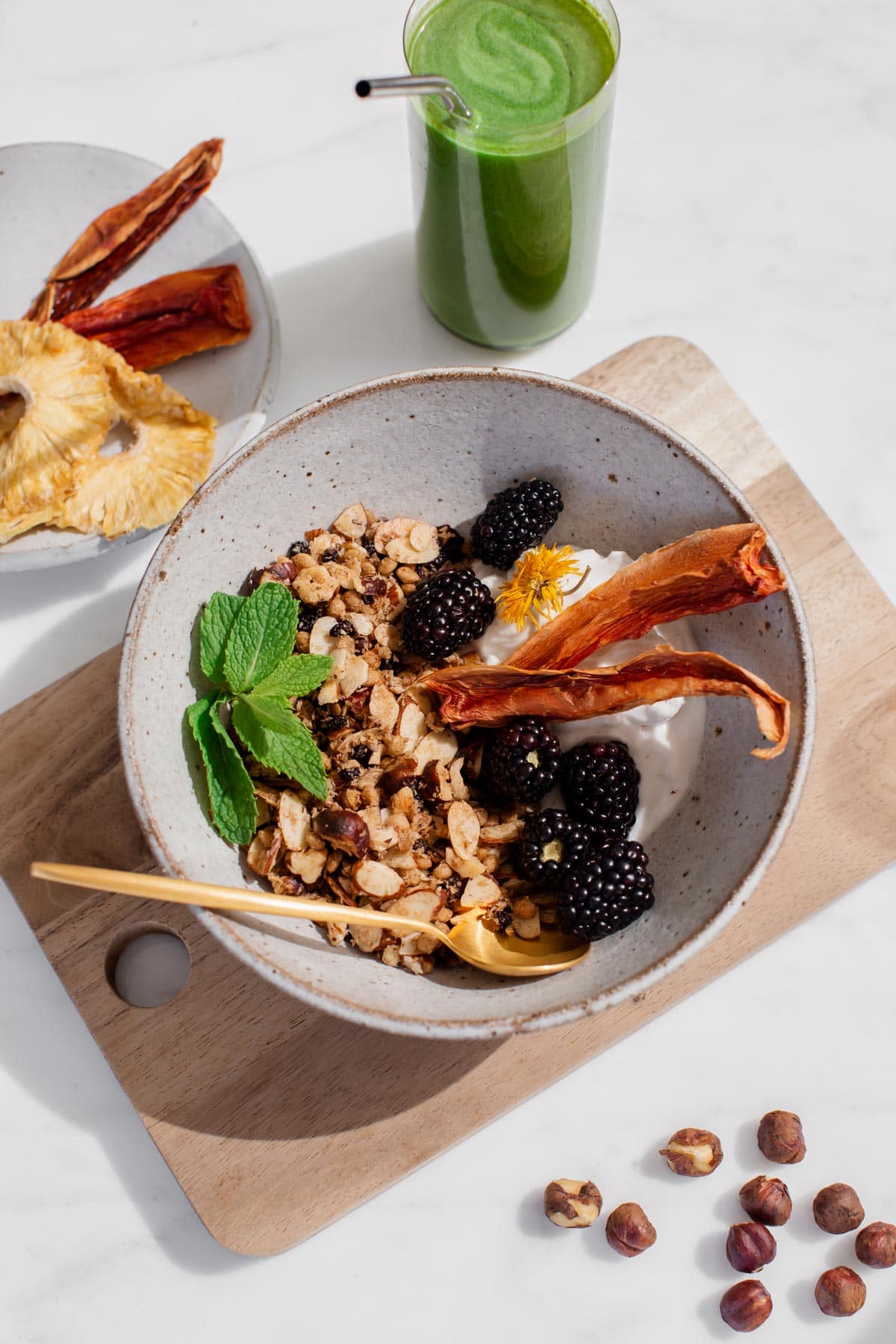 Hero shot of a bowl of hazelnut granola topped with blackberries, papaya and a sprig of mint, on a wooden board, with a gold spoon and vivid green juice beside it.