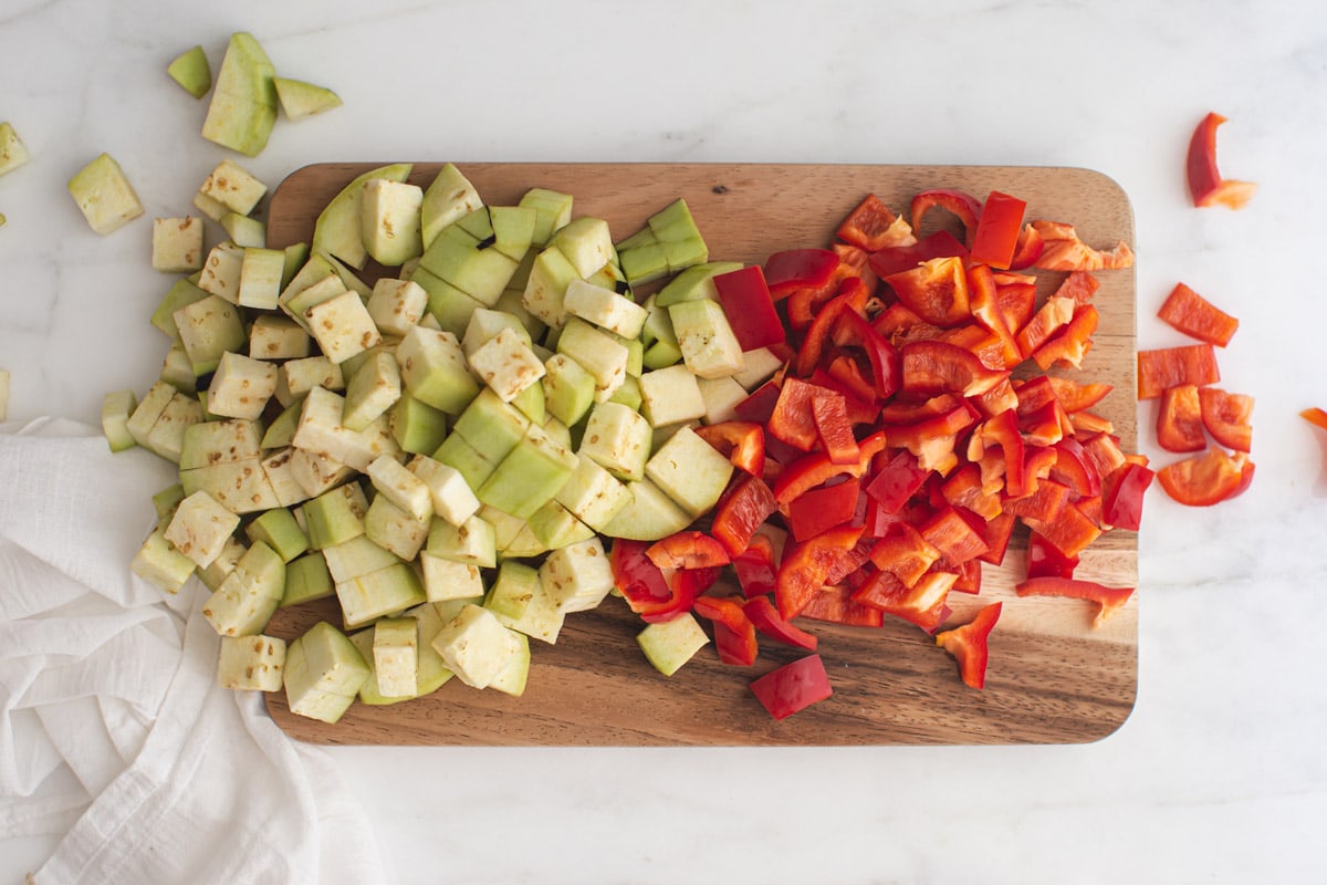 Wooden chopping board piled with cubed eggplant and sliced bell peppers