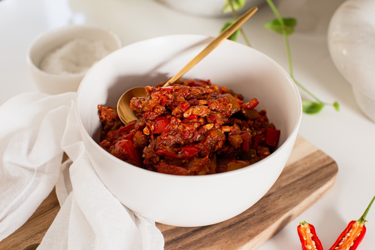 Bowl of cooked eggplant and bell pepper dip on a wooden serving board, with fresh chili in the background.