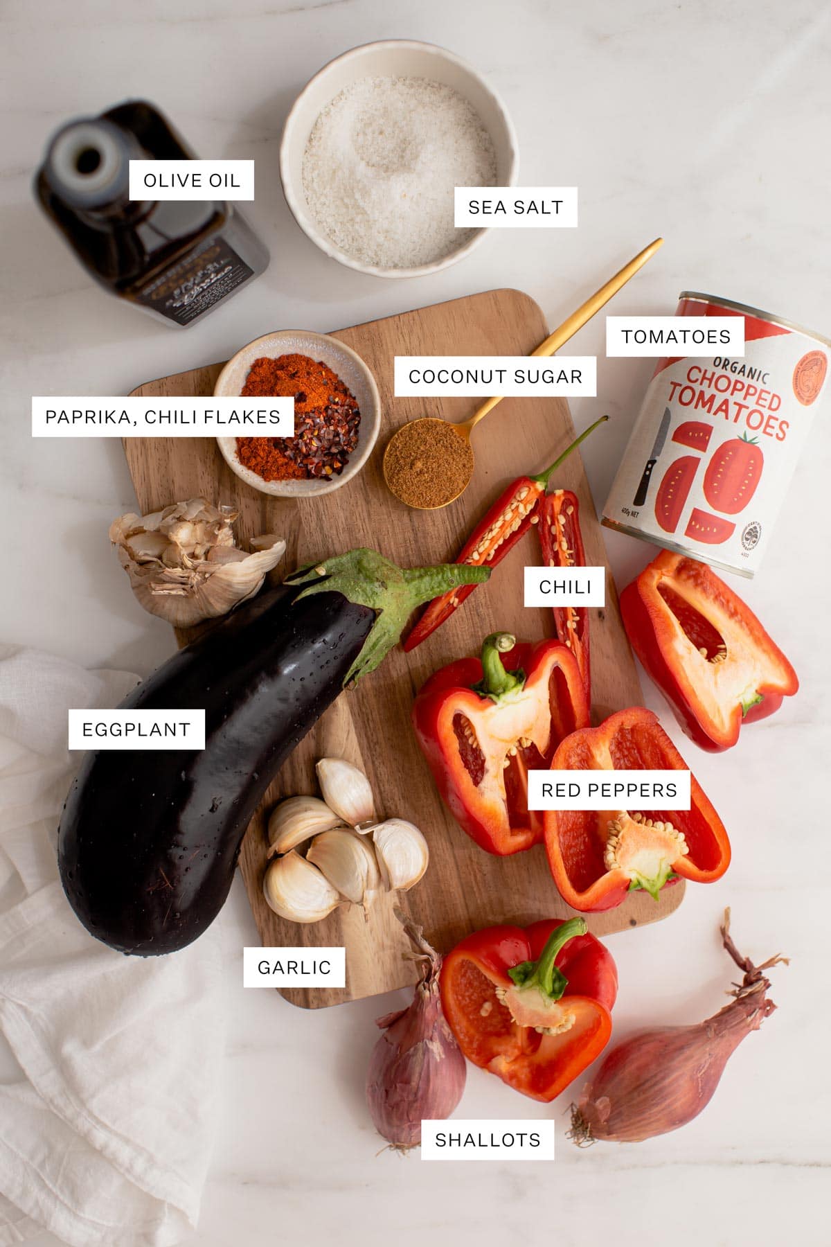 Flat lay of all the ingredients used to make this recipe - olive oil, salt, paprika, chili, coconut sugar, tinned tomatoes, eggplant, bell peppers and garlic.