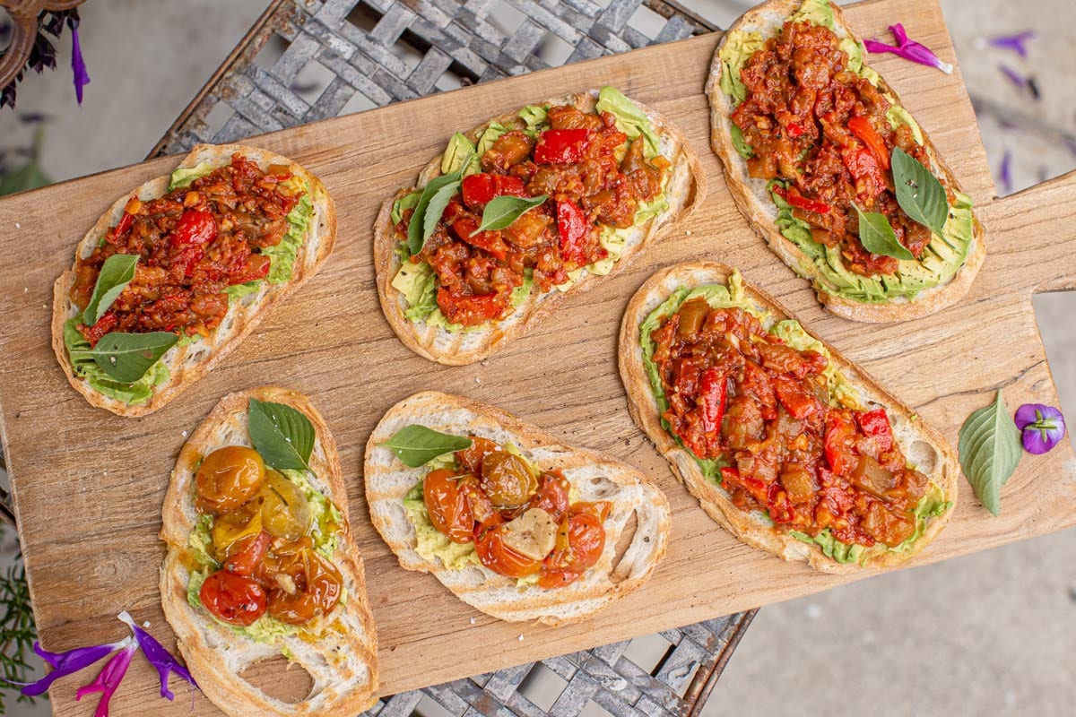 Wooden serving board on a rustic outdoor table surrounded by rosemary and sage flowers. On the board are slices of toasted sourdough topped with matbucha, they look rich and vibrant