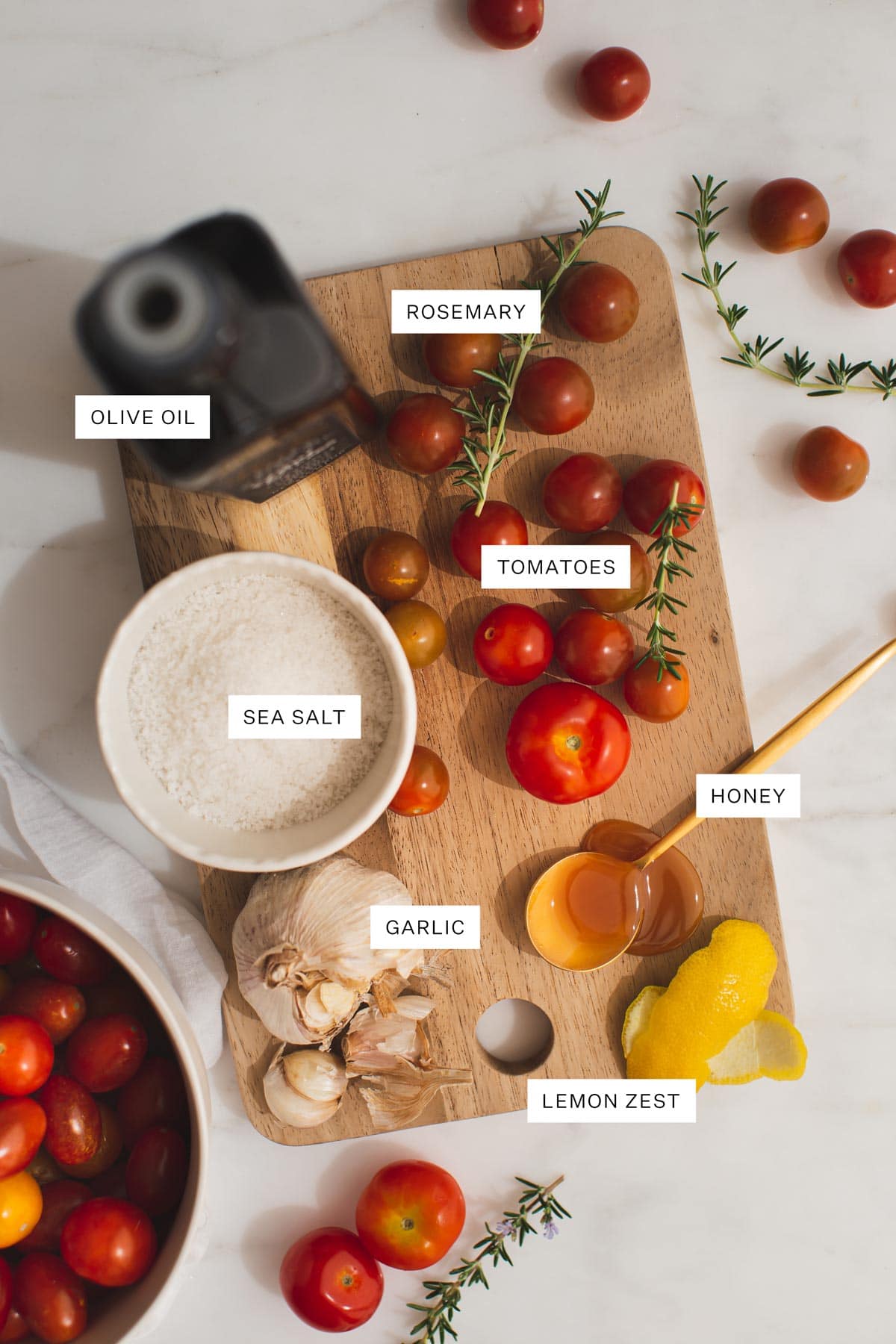 Flat lay of ingredients needed to make this recipe, including quality olive oil, cherry tomatoes, lemon, fresh rosemary, sea salt, pepper, honey.