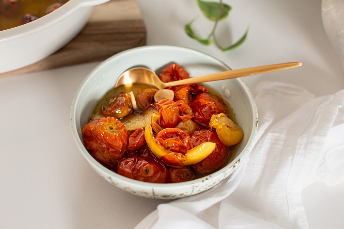 A ceramic bowl filled with confit tomatoes, sliced garlic and lemon peel, on a wooden serving board