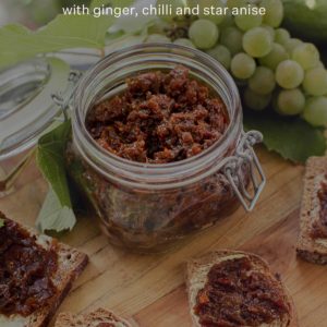 A jar of homemade feijoa chutney on a wooden board surrounded by fresh grapes and rye sourdough. Text over the top says "Feijoa Chutney with ginger, chilli and star anise".