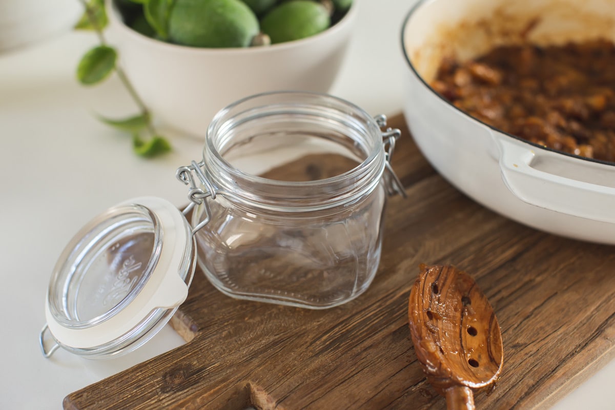 A small glass preserving jar with a rubber seal on a wooden board, beside a saucepan filled with cooked chutney