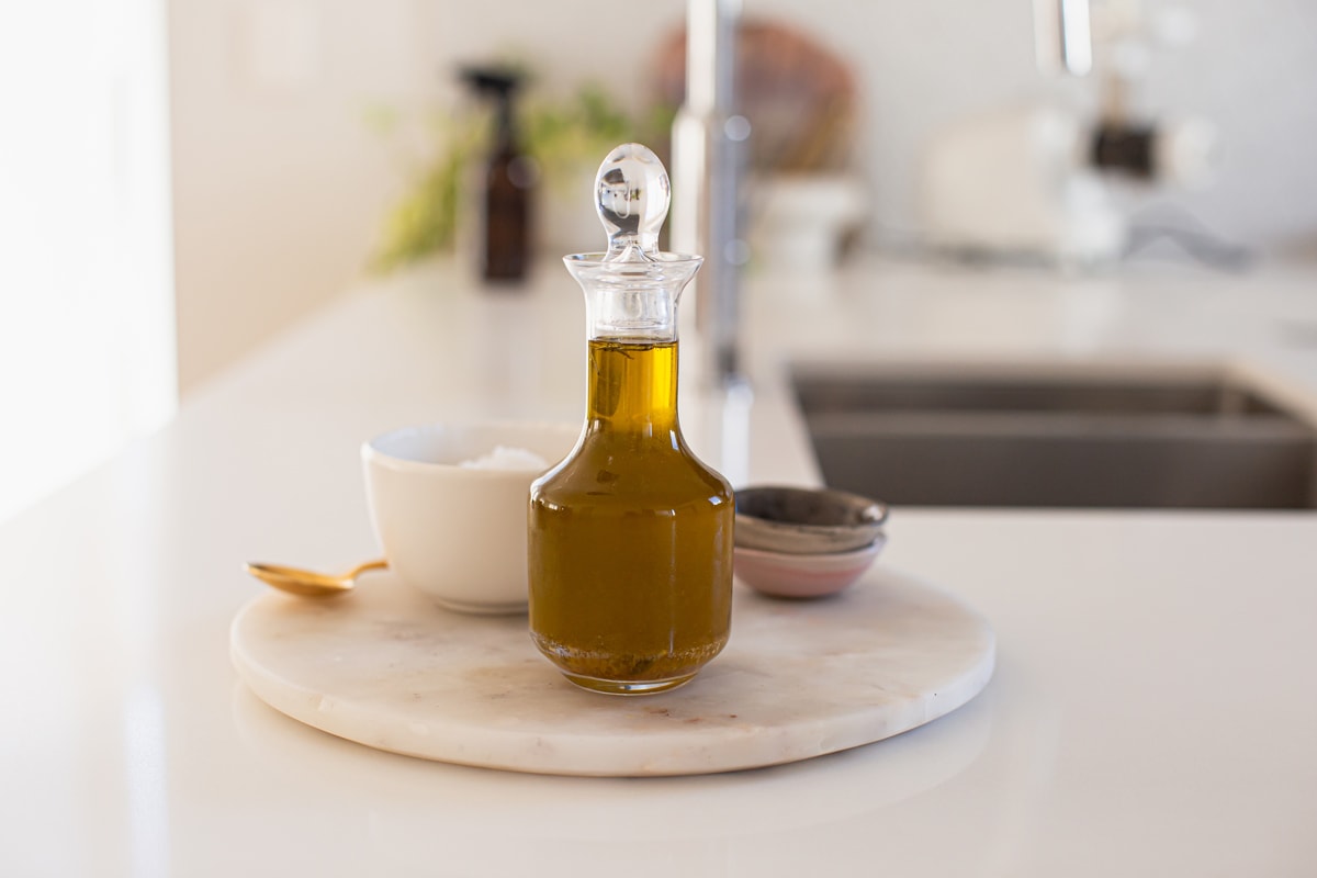 A glass bottle with a stopper filled with the remaining confit oil, on a marble platter beside other seasonings - salt, black pepper.