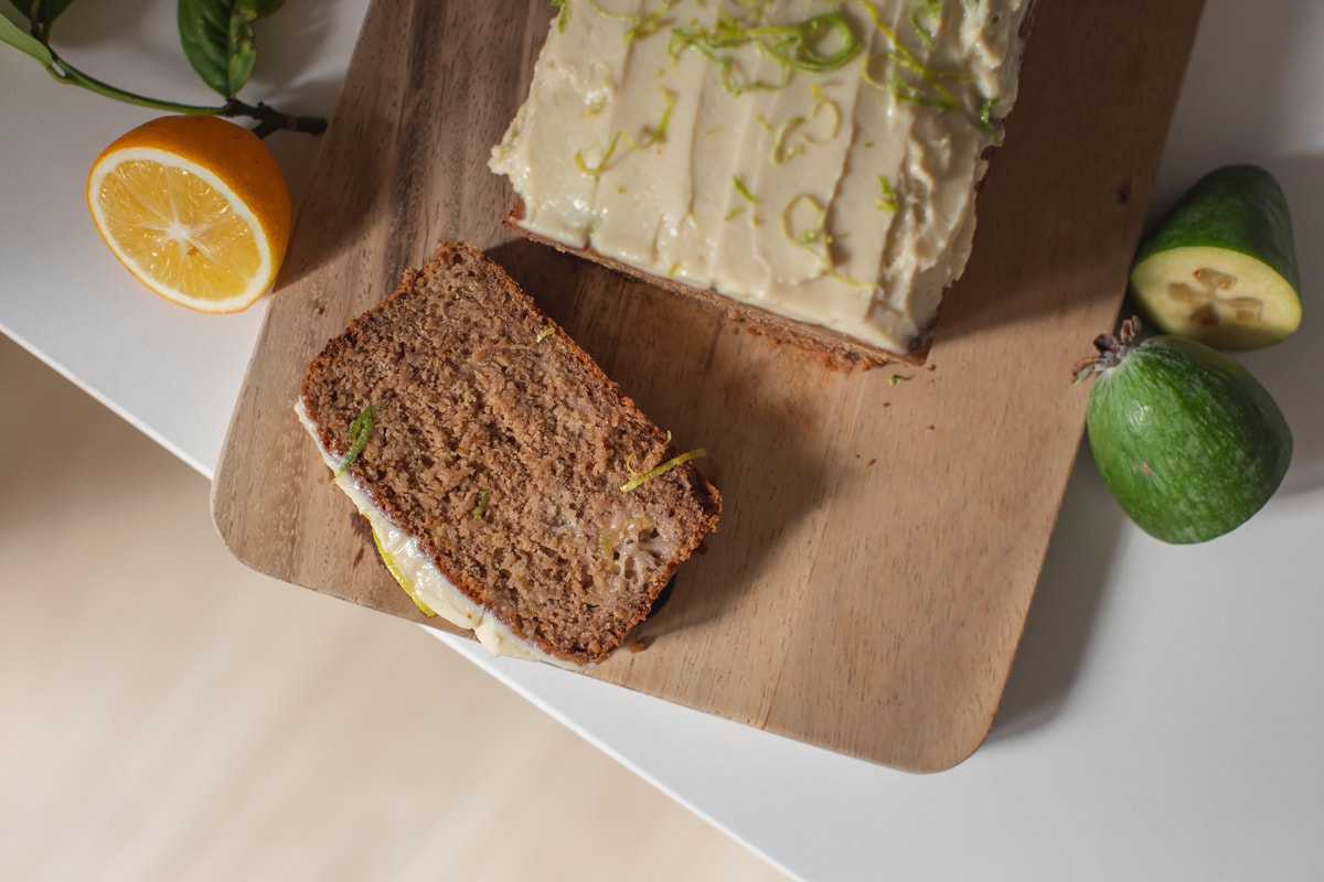 Overhead shot of the freshly baked loaf, with a slice resting on a wooden board. The loaf is on the kitchen bench and surrounded by fresh feijoas and lemon.