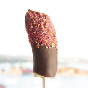 Close up of a banana pop coated in chocolate and dipped in crushed nuts and freeze dried fruit powder.