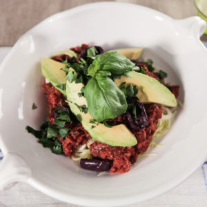 Bowl of zucchini pasta topped with marinara sauce, avocado, black olives and fresh herbs.