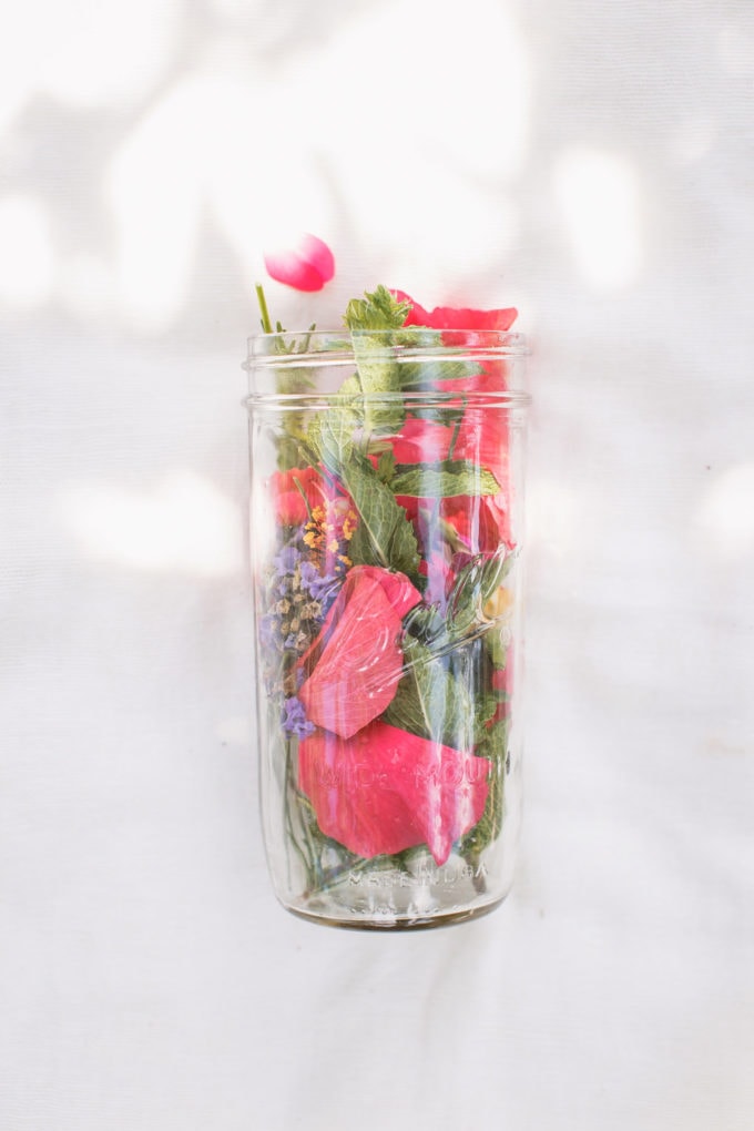 A large mason jar filled to the brim with colourful herbs and flowers, laying on a cream linen towel, dappled with light.