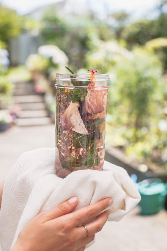 Lauren holding a large glass jar of water, filled with colourful herbs and flowers, it's freshly made moon water. A garden scene is in the background.