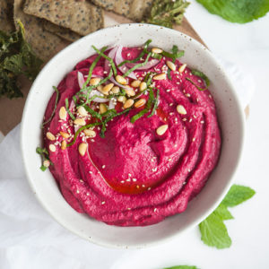 A ceramic bowl filled with vibrant pink roasted beet hummus, with toasted pine nuts and fresh herbs on top.