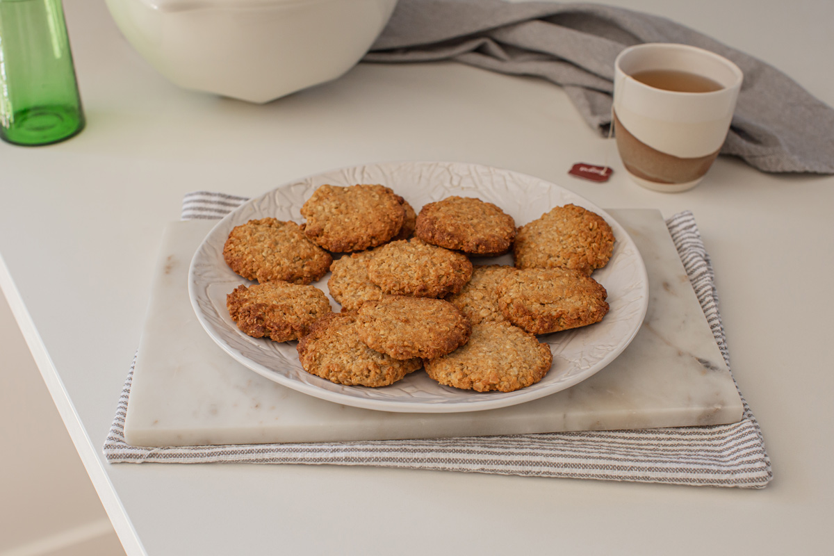 A plate of freshly baked vegan ANZAC biscuits rests on a marble board ready to enjoy. There is a tea towel and mug of tea in the background.