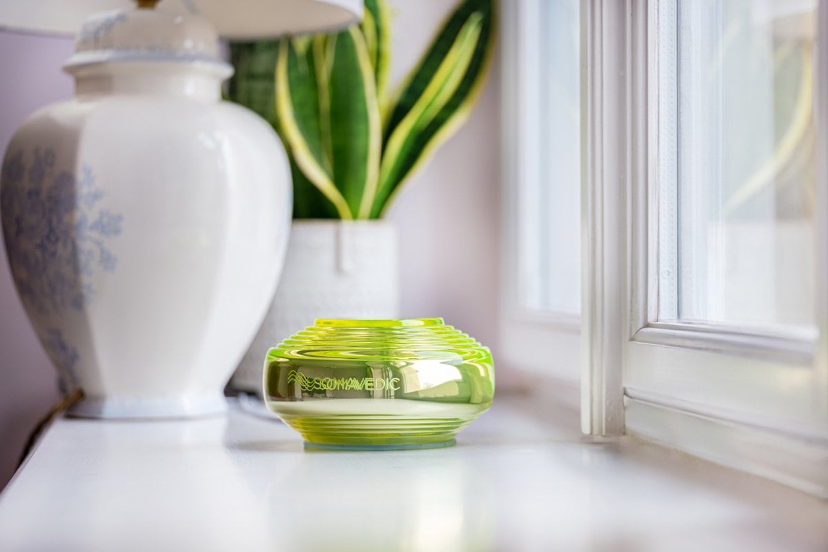 The EMF protective Somavedic device sitting on a cabinet bby the window with a plant in the background.