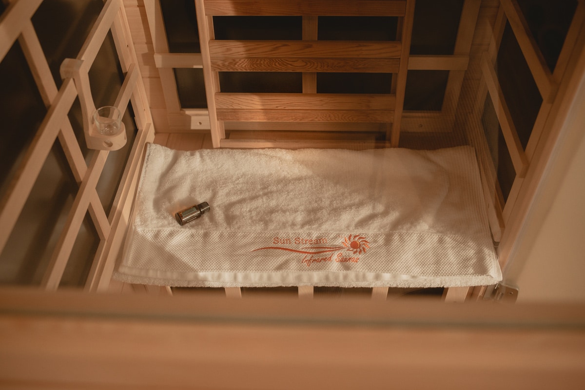 View of the inside of a small home infrared sauna, showing a fluffy towel on the bench and sunlight streaming in.