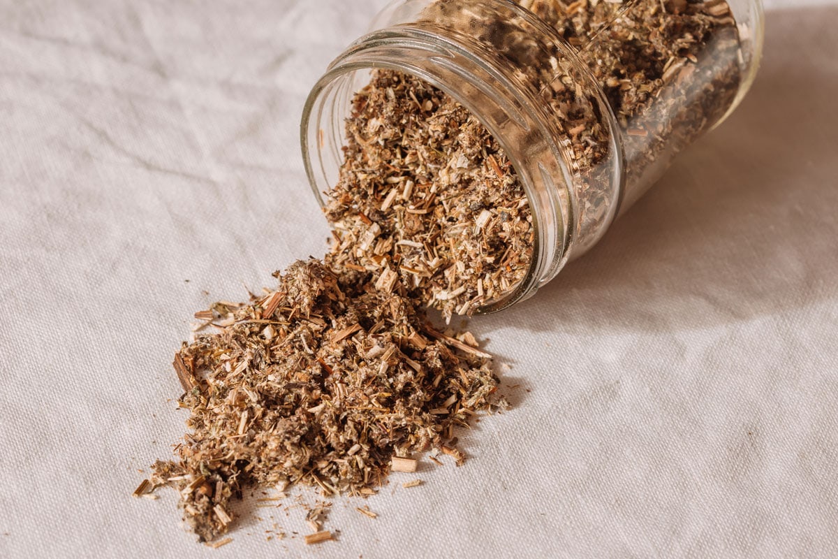 A glass jar laying sideways on a kitchen table, with dried mugwort leaves spilling out over the table linen.