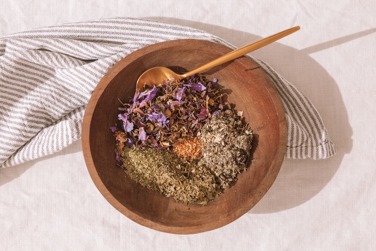 A wooden bowl on a table covered in linen, filled with a variety of herbs used to make this tea blend.