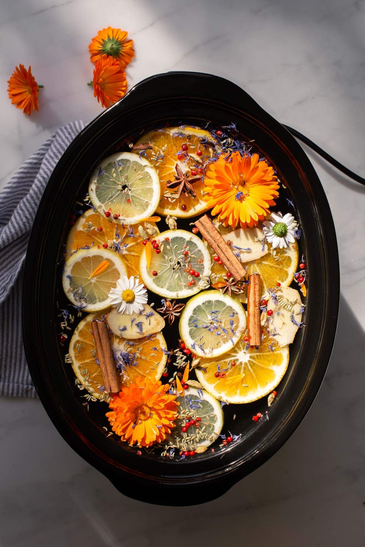 Hero shot of a crockpot on a marble counter, filled with the most vibrant colourful ingredients: sliced orange, grapefruit and limes, pink peppercorns, star anise, cinnamon sticks, orange calendula flowers, purple cornflowers and more. The lid is off, to allow the scent to fill the home.