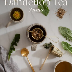 Mugs of dandelion tea on a kitchen counter, with leaves and flower heads strewn about. Text over the top reads; "How to make dandelion tea, 3 ways."