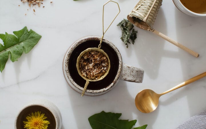 A mug of roasted dandelion tea with a brass tea strainer resting over the top. Dandelion leaves and petals are strewn about over the marble counter.